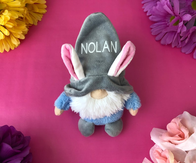 Puede incluir: Un gnomo de peluche gris con una camisa azul y barba blanca que lleva un gorro de conejo gris con orejas rosas. El gorro tiene el nombre "NOLAN" bordado.