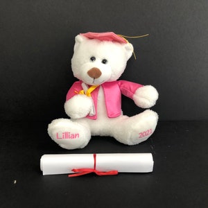 Pu&ograve; includere: Un orsacchiotto bianco che indossa una toga da laurea rosa e un cappello. L'orso tiene un rotolo giallo e ha il nome "Lillian" su un piede e "2021" sull'altro. Un diploma bianco &egrave; arrotolato davanti all'orso.
