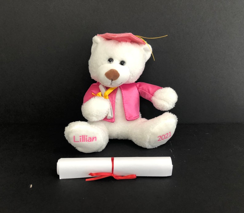 Pu&ograve; includere: Un orsacchiotto bianco che indossa una toga da laurea rosa e un cappello. L'orso tiene un rotolo giallo e ha il nome "Lillian" su un piede e "2021" sull'altro. Un diploma bianco &egrave; arrotolato davanti all'orso.