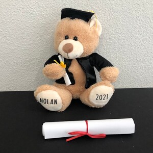 Pu&ograve; includere: Un orsacchiotto di peluche marrone che indossa una toga da laurea nera e un cappello, tenendo un diploma. L'orso ha il nome "NOLAN" e l'anno "2021" ricamato sulle zampe.