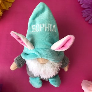 Puede incluir: Un gnomo de peluche azul claro con orejas de conejo rosas y un nombre bordado en blanco "Sophia" en el gorro. El gnomo lleva una barba peluda gris y blanca y est&aacute; sentado sobre un fondo rosa.