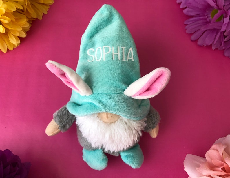 Puede incluir: Un gnomo de peluche azul claro con orejas de conejo rosas y un nombre bordado en blanco "Sophia" en el gorro. El gnomo lleva una barba peluda gris y blanca y est&aacute; sentado sobre un fondo rosa.
