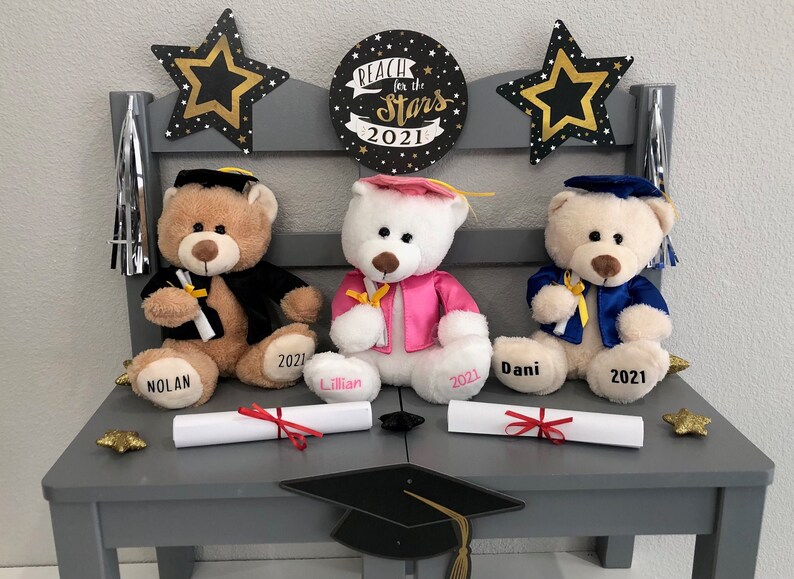 Pu&ograve; includere: Tre orsacchiotti vestiti con toge e cappelli da laurea, seduti su una panchina grigia con un cartello che recita "Reach for the Stars 2021". Ogni orsacchiotto tiene un diploma e un cartellino con l'anno 2021.