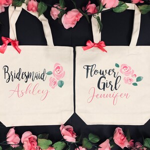 Puede incluir: Dos bolsas de lona blancas con dise&ntilde;os florales rosas y texto negro. La bolsa de la izquierda dice "Bridesmaid Ashley" y la bolsa de la derecha dice "Flower Girl Jennifer".