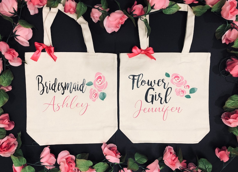 Puede incluir: Dos bolsas de lona blancas con dise&ntilde;os florales rosas y texto negro. La bolsa de la izquierda dice "Bridesmaid Ashley" y la bolsa de la derecha dice "Flower Girl Jennifer".