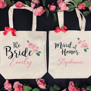 Puede incluir: Dos bolsas de lona blancas con dise&ntilde;os florales rosas y asas de cinta rosa. La bolsa de la izquierda dice "The Bride Emily" y la bolsa de la derecha dice "Maid of Honor Stephanie".