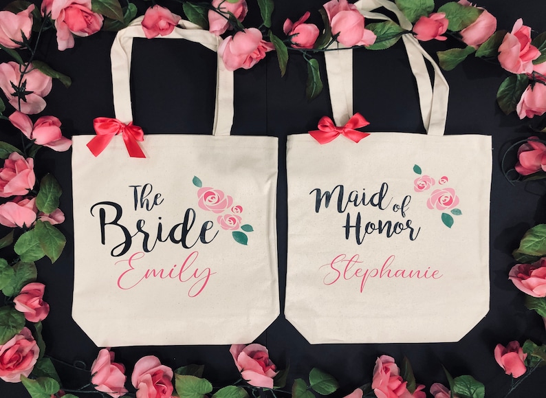 Puede incluir: Dos bolsas de lona blancas con dise&ntilde;os florales rosas y asas de cinta rosa. La bolsa de la izquierda dice "The Bride Emily" y la bolsa de la derecha dice "Maid of Honor Stephanie".