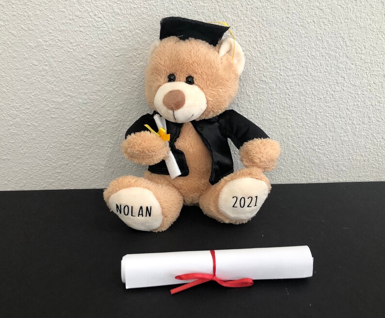 Pu&ograve; includere: Un orsacchiotto di peluche marrone che indossa una toga da laurea nera e un cappello. L'orso tiene un diploma e ha il nome "Nolan" e l'anno "2021" ricamato sulle zampe.