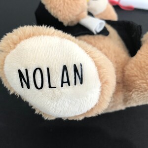 Pu&ograve; includere: Un orsacchiotto di peluche marrone che indossa una toga da laurea nera e un cappello. Il piede dell'orso ha il nome "NOLAN" ricamato.