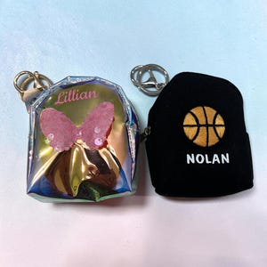 May include: Two miniature backpack keychains. One is iridescent silver with a pink sequined butterfly and the name Lillian printed in pink. The other is black fabric with an embroidered basketball and the name NOLAN printed in white text.