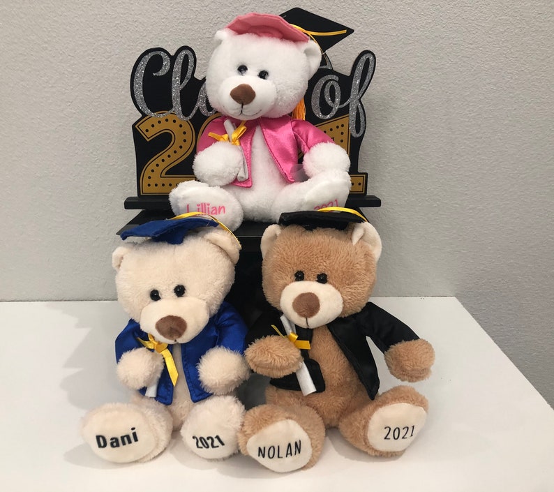 Pu&ograve; includere: Tre orsacchiotti che indossano cappelli e toge da laurea. Gli orsi sono seduti su un cartello di legno nero che dice "Classe del 2021". Gli orsi si chiamano Dani, Nolan e Lillian.