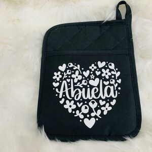 May include: Black quilted oven mitt with a white heart design. The heart features the word "Abuela" surrounded by flowers and small hearts.