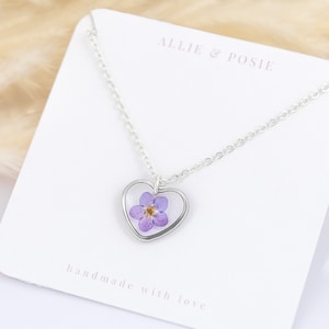 May include: A silver chain necklace with a heart-shaped pendant featuring a pressed purple flower. The pendant is surrounded by a silver frame. The card reads "Allie & Posie" and "handmade with love".