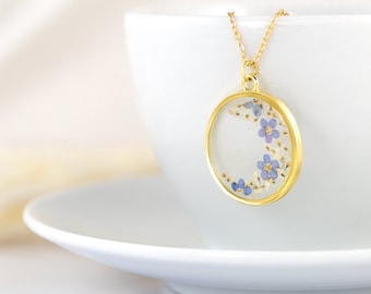 May include: A gold necklace with a round pendant featuring a crescent moon shape filled with pressed forget-me-not flowers.