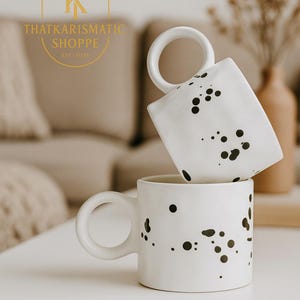 May include: Two white ceramic mugs with large, rounded handles are stacked on a white surface. Each mug is decorated with irregular black splatters. The mugs are part of the That Karismatic Shoppe collection, established in 2024.