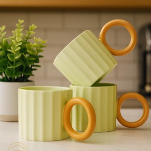 May include: Three light green ceramic mugs with ribbed sides and orange ring handles are stacked on a white surface. A small potted plant is in the background. The mugs have the text "THATKARISMATIC SHOPPE" on the bottom.