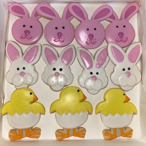 May include: A box of Easter-themed sugar cookies. There are nine cookies in total: six bunny cookies, three chick cookies. The bunny cookies are pink and white with icing details. The chick cookies are yellow with white icing and orange feet.