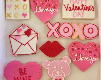 May include: A box of nine Valentine's Day sugar cookies decorated with pink, red, and gold icing. The cookies feature various designs, including hearts, XOXO, a teddy bear, a letter, and the phrases "Happy Valentine's Day", "I love you", and "Be Mine".