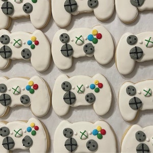 Może przedstawiać: Kolekcja białych i szarych ciasteczek cukrowych ozdobionych kolorowymi guzikami i zielonym X, w kształcie kontrolera do gier wideo.
