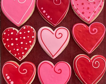 May include: Nine heart-shaped sugar cookies decorated with pink and red icing. Some cookies have white polka dots, while others have a swirl design.