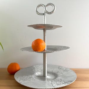 May include: A three-tiered silver serving tray with an infinity symbol handle. The tray has a floral design on the bottom tier and holds an orange. The tray is approximately 30 cm tall.