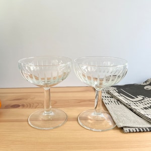 May include: Two clear glass champagne coupes with a fluted design. The glasses are empty and sitting on a wooden surface.