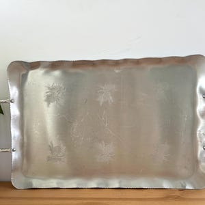 May include: A rectangular silver serving tray with handles. The tray has a scalloped edge and embossed floral designs. The handles are attached with rivets. The tray is on a wooden surface.