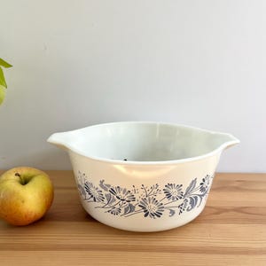 May include: Cream-colored vintage mixing bowl with a floral design in shades of gray and blue. The bowl has a white interior and two handles. A yellow and red apple sits to the left of the bowl on a wooden surface.