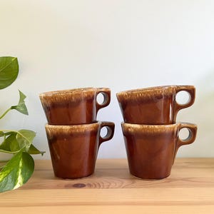 May include: Four brown ceramic mugs with a distinctive angled rim and a rounded handle. The mugs have a glossy finish with a lighter brown rim. They are stacked in pairs on a wooden surface.