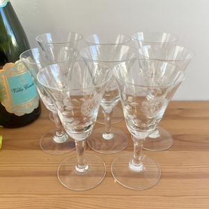May include: A collection of six clear, etched crystal wine glasses featuring floral patterns. The glasses have a slender stem and a wide, flared bowl. A green bottle of sparkling wine is partially visible in the background, with some greenery.