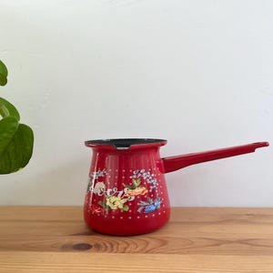 May include: A red enamel coffee pot with a long handle and floral design. The pot has a black rim and is decorated with colourful flowers and leaves. It sits on a wooden surface with a green plant in the background.