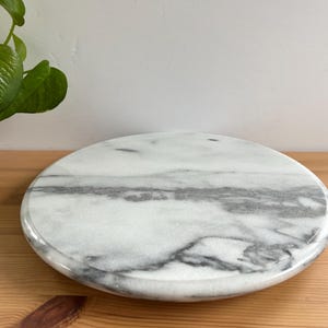 May include: A circular marble serving board with grey veining. The board is placed on a wooden surface, with a green plant in the background. This decorative board is ideal for serving cheese, appetizers, or desserts.