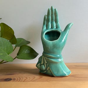 May include: A glossy turquoise ceramic vase shaped like an upright hand with fingers extended. The base of the hand features a draped fabric detail with a small circular accent. The palm area contains a hollow opening for holding small items.