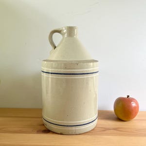 May include: A beige stoneware jug with a handle and a conical top, adorned with two blue stripes. A red and yellow apple sits to the right of the jug on a wooden surface. The jug is a vintage kitchenware item.