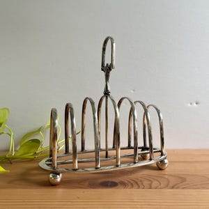 Peut inclure: Un porte-toast en métal argenté doté de six fentes arquées pour maintenir des tranches de pain. Le support est muni d'une poignée verticale centrale pour le transport et repose sur quatre petits pieds sphériques à la base.