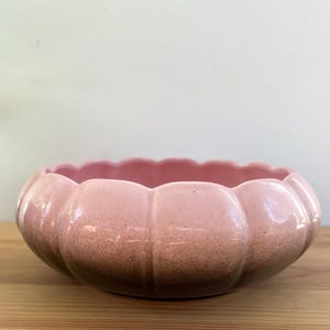 May include: A pink ceramic bowl with a scalloped rim and a pumpkin-like shape. The bowl has a speckled texture and a gradient colour, transitioning from a lighter pink to a darker brown at the base. It sits on a wooden surface.