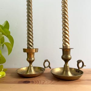 Peut inclure: Deux bougeoirs en laiton avec des bougies torsadées dorées. Les bougeoirs ont une base ronde, une petite poignée et un design détaillé. Les bougies sont de couleur or clair et ont un motif en spirale.