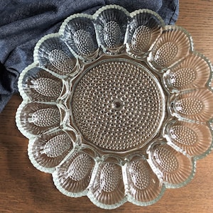 May include: Clear glass devilled egg serving platter with a scalloped edge. The centre and egg-shaped compartments have a textured, dotted pattern. The platter is on a wooden surface with a dark blue cloth.
