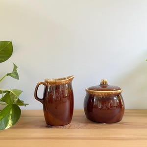 May include: A brown ceramic jug and lidded sugar bowl. The jug has a handle and a spout, and the sugar bowl has a lid with a small knob. Both items have a glossy finish and a two-tone colour scheme.