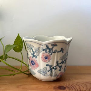 May include: A white ceramic planter with a scalloped rim and a fluted body. The exterior features a hand-painted pattern of pink flowers with blue centres, green leaves, and dark blue winding vines against a smooth, glossy white background.