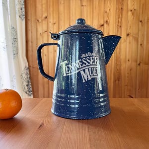 May include: A navy blue enamel coffee pot with white speckles and the text "Jack Daniel's Tennessee Mud". The pot has a lid, handle, and spout. Two oranges sit on a wooden table in front of the pot.
