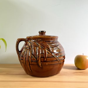 May include: A brown ceramic lidded pot with a handle and leaf design. The pot is on a wooden surface next to a green plant and a red and yellow apple.