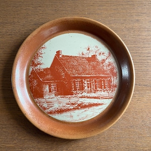 May include: Circular serving tray featuring a reddish-orange illustration of a house and trees on a white background. The tray has a brown rim, possibly made of wood. The tray is displayed on a wooden surface.