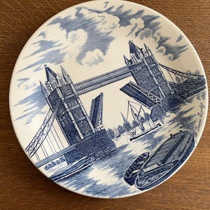 May include: A white ceramic plate with a blue illustration of Tower Bridge in London. The plate depicts the bridge, boats, and clouds. The words "Tower Bridge" are written on the right side of the plate.