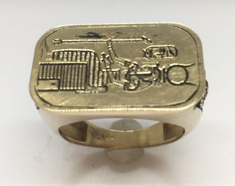 May include: A gold-toned ring with a rectangular design featuring an engraved image of a tractor. The ring has a smooth, polished band.