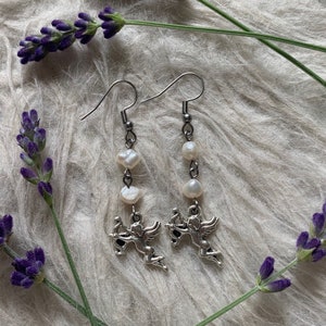 Puede incluir: Pendientes colgantes plateados con un peque&ntilde;o querub&iacute;n con arco y flecha, combinados con perlas blancas. Los pendientes se presentan sobre un fondo blanco suave, con ramitas de lavanda morada.