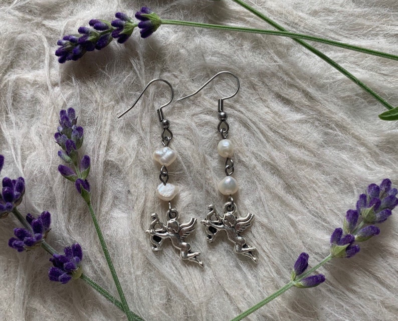 Puede incluir: Pendientes colgantes plateados con un peque&ntilde;o querub&iacute;n con arco y flecha, combinados con perlas blancas. Los pendientes se presentan sobre un fondo blanco suave, con ramitas de lavanda morada.