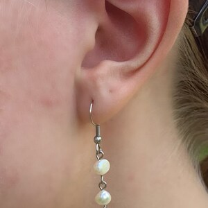 Puede incluir: Un pendiente colgante con un gancho plateado, dos perlas blancas y un peque&ntilde;o cupido plateado con arco y flecha. El pendiente se muestra en la oreja de una persona.