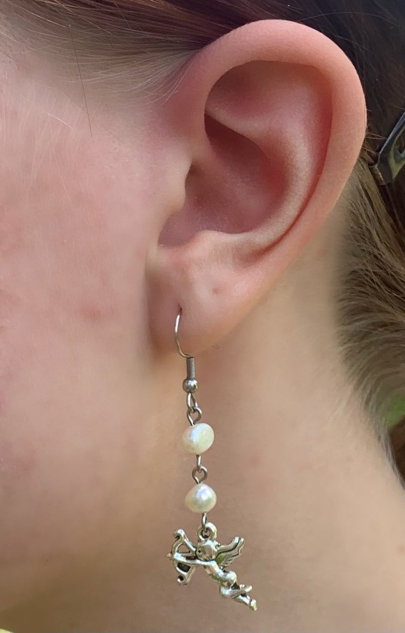 Puede incluir: Un pendiente colgante con un gancho plateado, dos perlas blancas y un peque&ntilde;o cupido plateado con arco y flecha. El pendiente se muestra en la oreja de una persona.