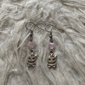 May include: A pair of silver-toned earrings. Each earring features a small, pink heart-shaped bead above a skull charm with a bow and crossbones. The earrings are suspended from a simple hook.
